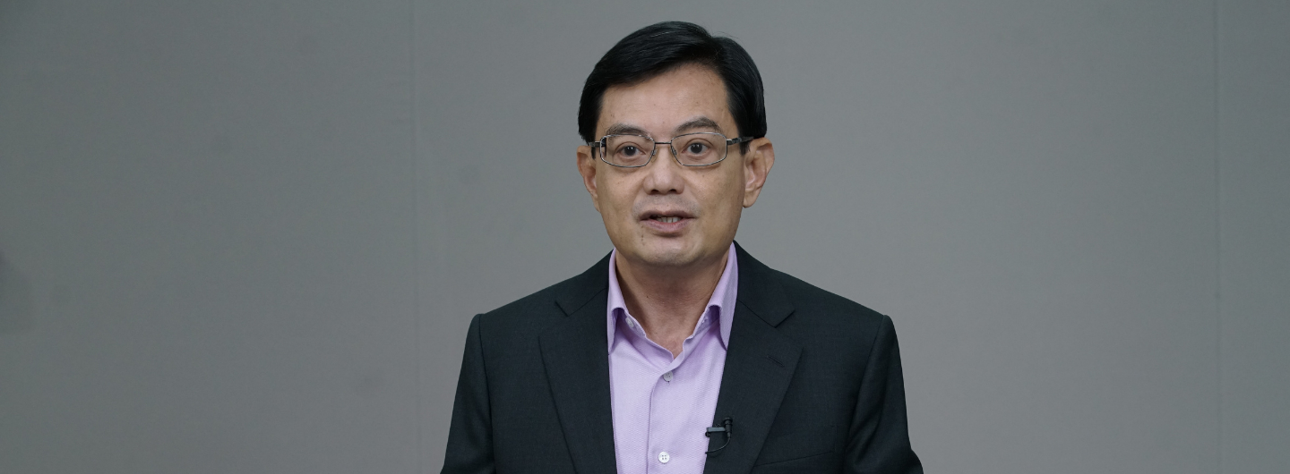 Man in suit and glasses, lilac shirt, speaking against plain gray background.